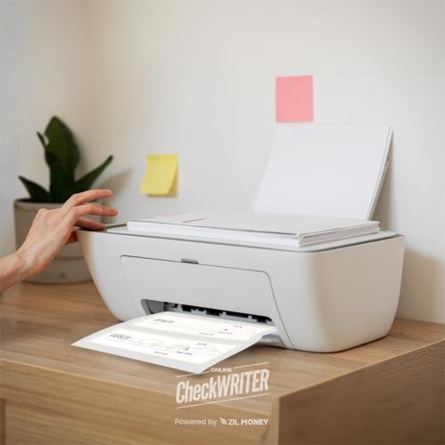 A Person Printing a Blank Check Print Document on a Compact White Printer Placed on a Wooden Desk, with Sticky Notes on the Wall for Reminders