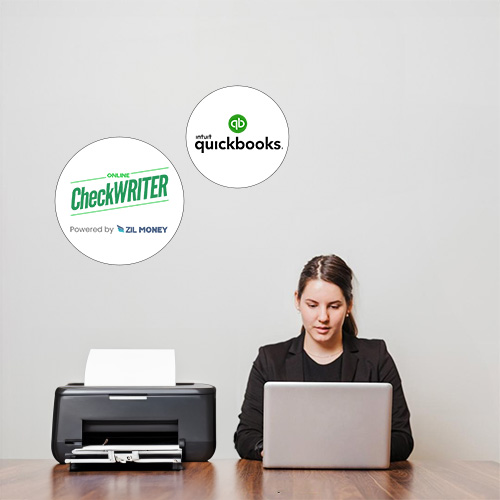 A Woman Sits at a Desk Using a Laptop with Checks For QuickBooks Printing Using a Printer, a Logo on the Wall Behind Indicates QuickBooks