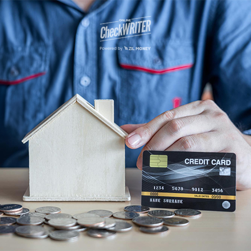At a Table, a Person in a Blue Shirt Holds a Credit Card Next to a Wooden House Model He Is Thinking About How to Pay Mortgage with Credit Card