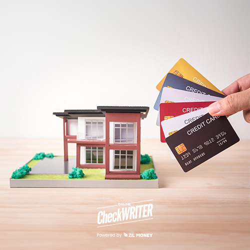 A Hand Holding Multiple Credit Cards Over a Model House on a Wooden Table Suggests Pay Your Mortgage With A Credit Card