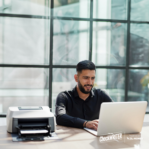 Focused Man Using Costco Checks Business Alternative on a Laptop Beside a Printer Printing Checks