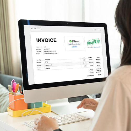 A Person Sits at a Desk Using a Computer. The Computer Screen Displays a QuickBooks Invoice Generator.