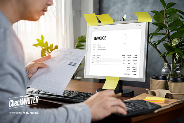 A Person Sits at a Desk Using a Computer. The Computer Screen Displays an Invoice Software