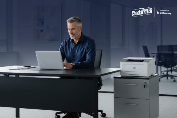 5 Reasons why Smart Business Owners choose to print checks themselves! Business owner working on a laptop beside a printer to Print Checks Yourself securely from the office.