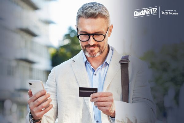 5 Reasons Paying Vendors by Credit Card Can Improve Cash Flow Timing Man in glasses holding a phone and card to Pay By Credit Card, highlighting secure on-the-go payments.